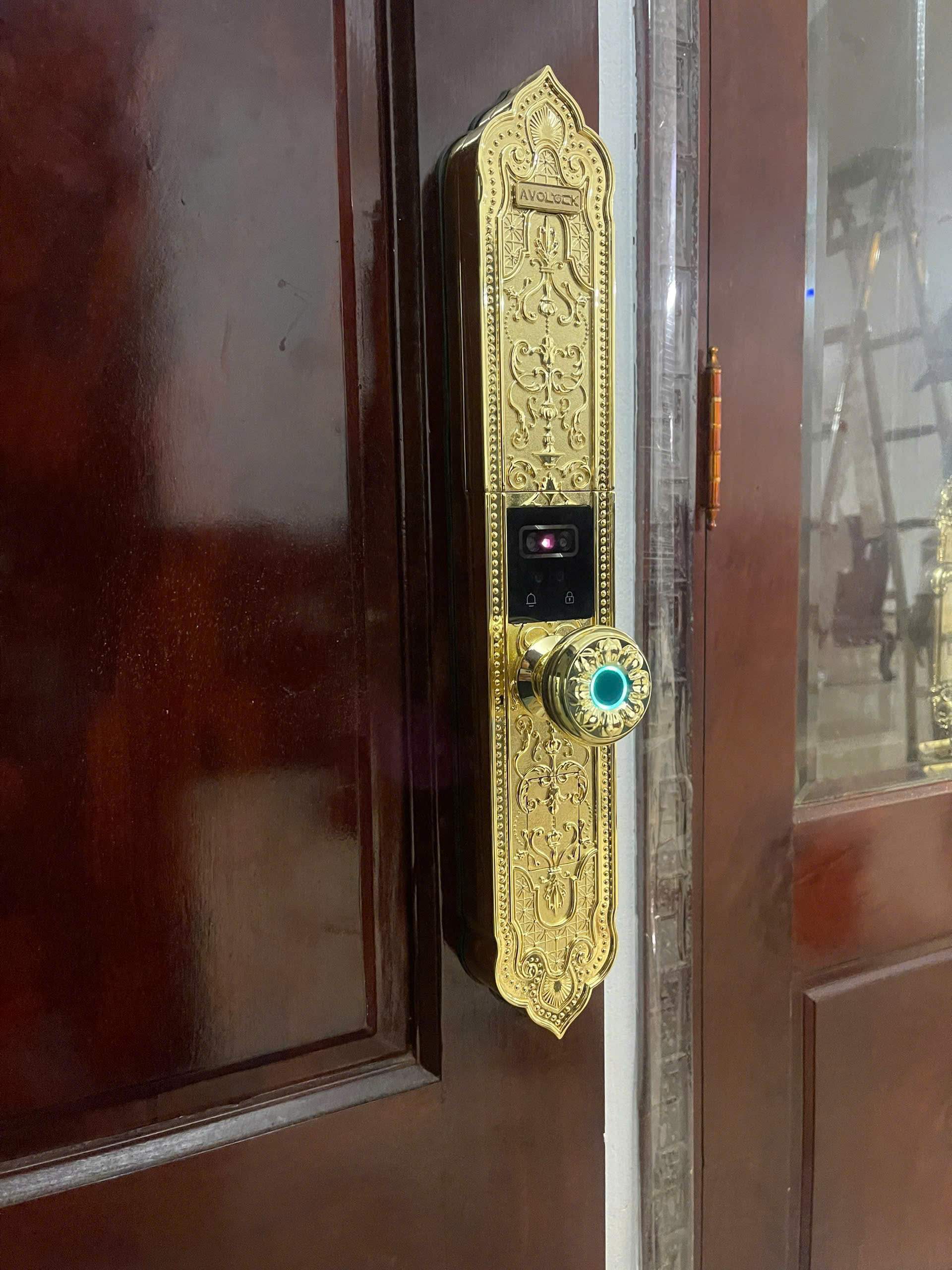 Cận cảnh mặt trước khóa Avolock AC-996F: họa tiết tân cổ điển mạ vàng, camera Face ID, núm vân tay sáng đèn xanh